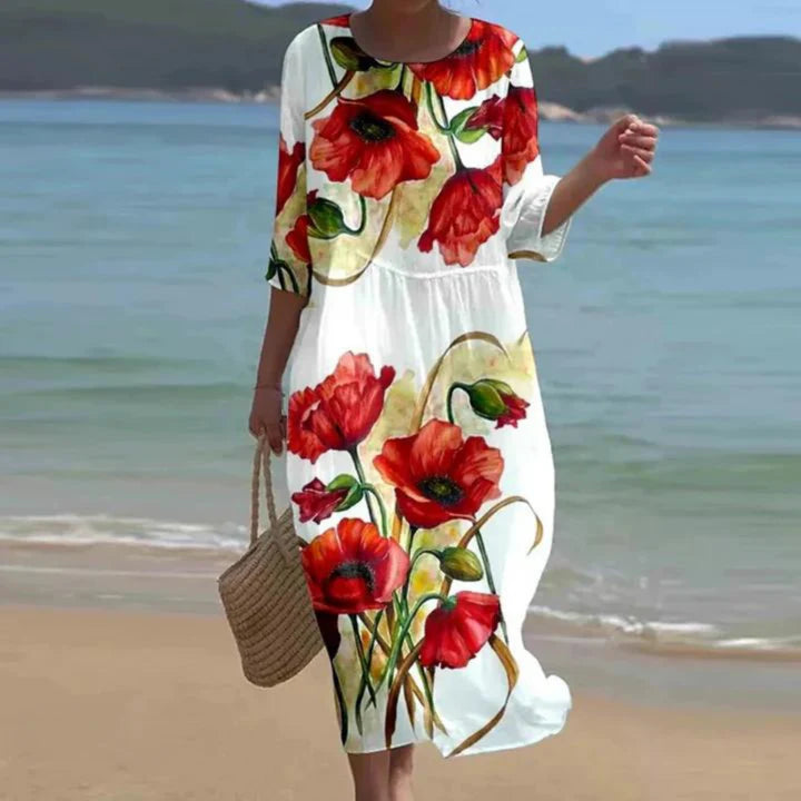 Frau in weißem Kleid mit rotem Mohnblumenmuster am Strand, trägt Strohtasche. Sommermode, Strandkleid, florales Design.