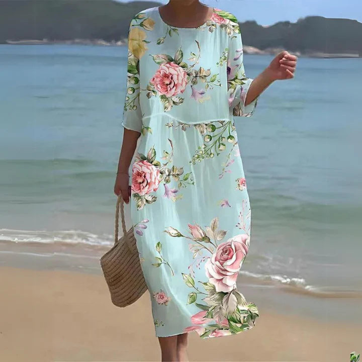 Frau am Strand in einem blumigen Sommerkleid mit Rosenmuster, trägt eine Strohtasche. Perfekt für Strandmode und Sommeroutfits.
