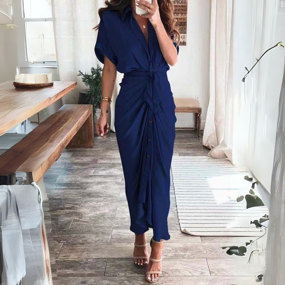 Frau in elegantem, blauem Wickelkleid, steht in modernem Wohnzimmer. Stilvolle Mode, Sommerkleid, Inneneinrichtung, Holztisch, helle Fenster.