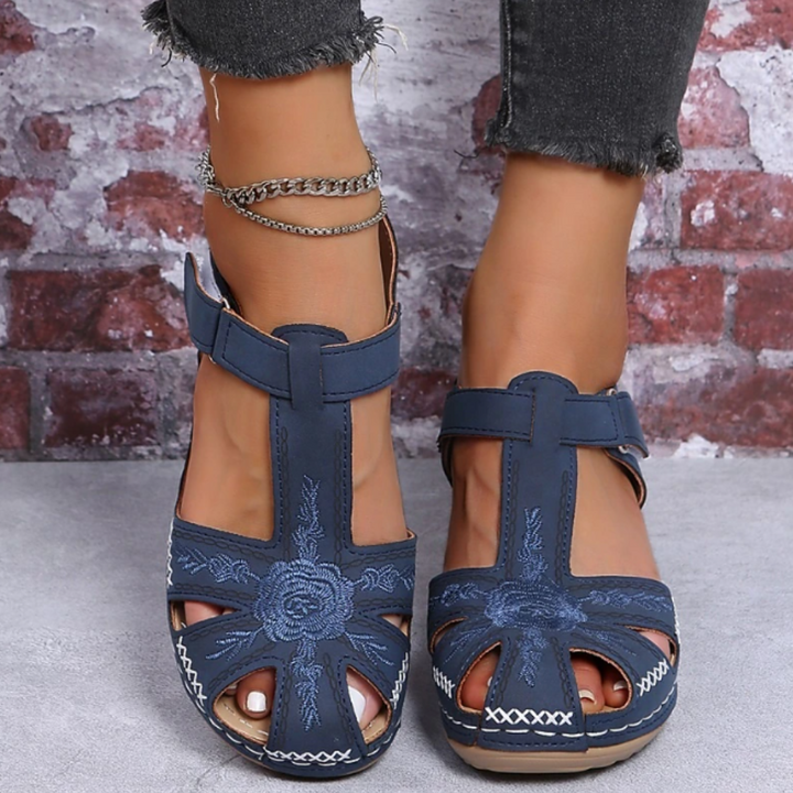 Damenfüße in blauen Sandalen mit Blumenstickerei, vor Ziegelwand. Modische Sommerschuhe, offene Zehen, Jeans mit Fransen, Fußkettchen.