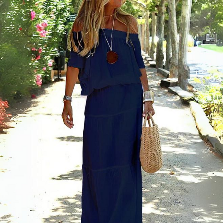 Frau in blauem Off-Shoulder-Kleid mit Strohtasche spaziert auf sonnigem Weg, umgeben von Bäumen und Blumen. Sommermode, Freizeitkleidung, Natur.