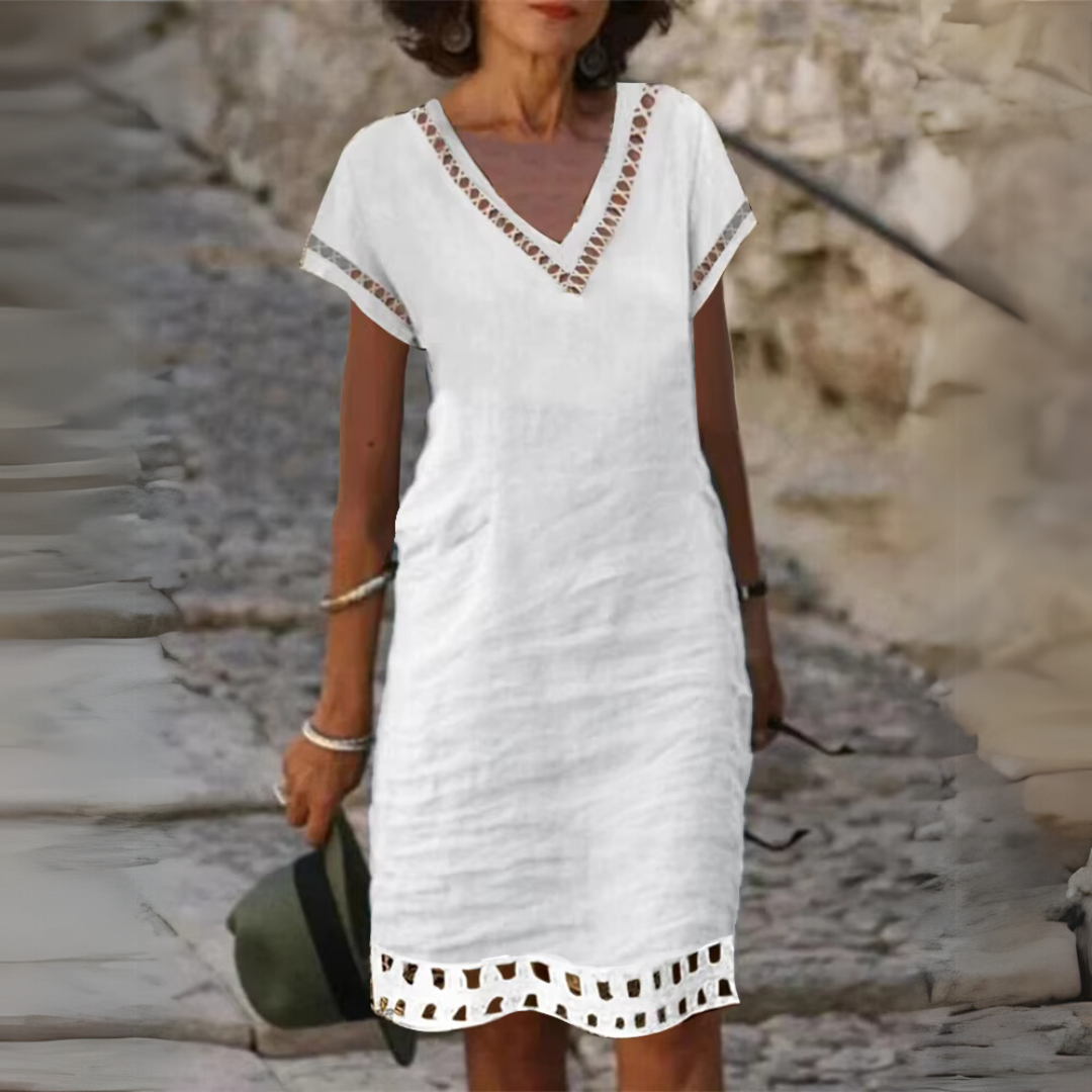 Frau in weißem Sommerkleid mit V-Ausschnitt und Lochmuster, spaziert auf Kopfsteinpflaster. Elegante Mode, lässiger Stil, Sommeroutfit.