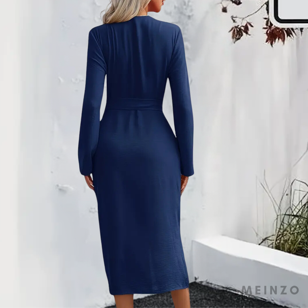 Frau in elegantem, langem, dunkelblauem Kleid mit langen Ärmeln, steht vor weißer Wand. Herbstliche Blätter im Hintergrund. Mode, Damenkleidung.