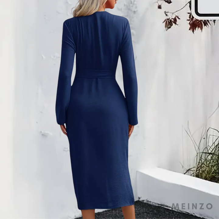 Frau in elegantem, langem, dunkelblauem Kleid mit langen Ärmeln, steht vor weißer Wand. Herbstliche Blätter im Hintergrund. Mode, Damenkleidung.