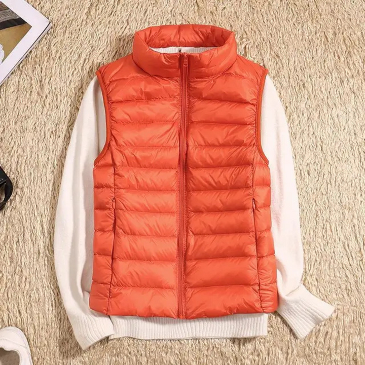 Orangefarbene Steppweste auf cremefarbenem Pullover, flach auf beigem Teppich liegend. Modetrend, Herbstmode, Damenbekleidung, warme Weste.