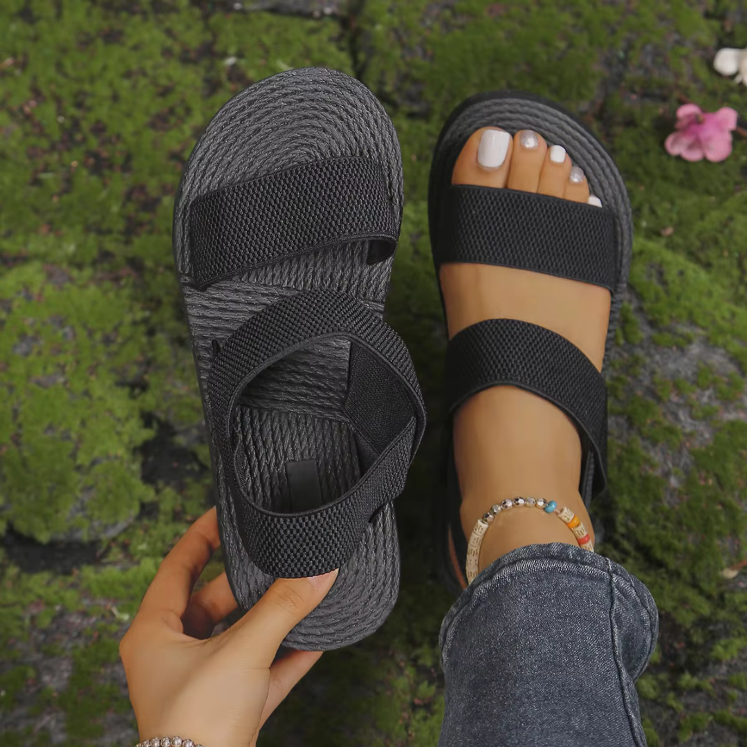 Schwarze Sandalen mit breiten Riemen auf moosbedecktem Boden, eine Sandale getragen, die andere in der Hand gehalten, modische Fußkettchen sichtbar.