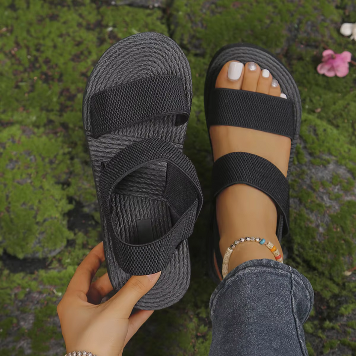 Schwarze Sandalen mit breiten Riemen auf moosbedecktem Boden, eine Sandale getragen, die andere in der Hand gehalten, modische Fußkettchen sichtbar.