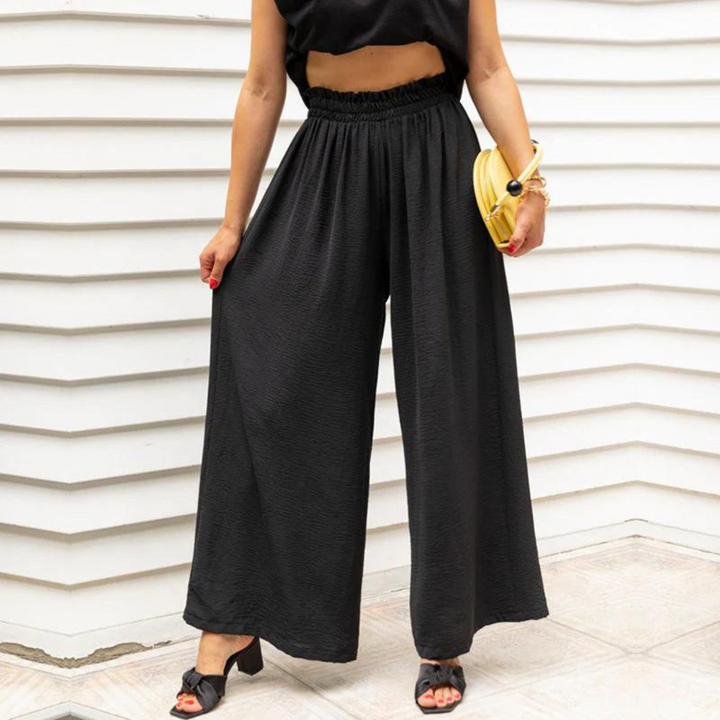 Frau in schwarzer, weiter Hose und schwarzem Top vor weißer Wand, hält gelbe Clutch. Modetrend, Sommeroutfit, elegante Damenmode.