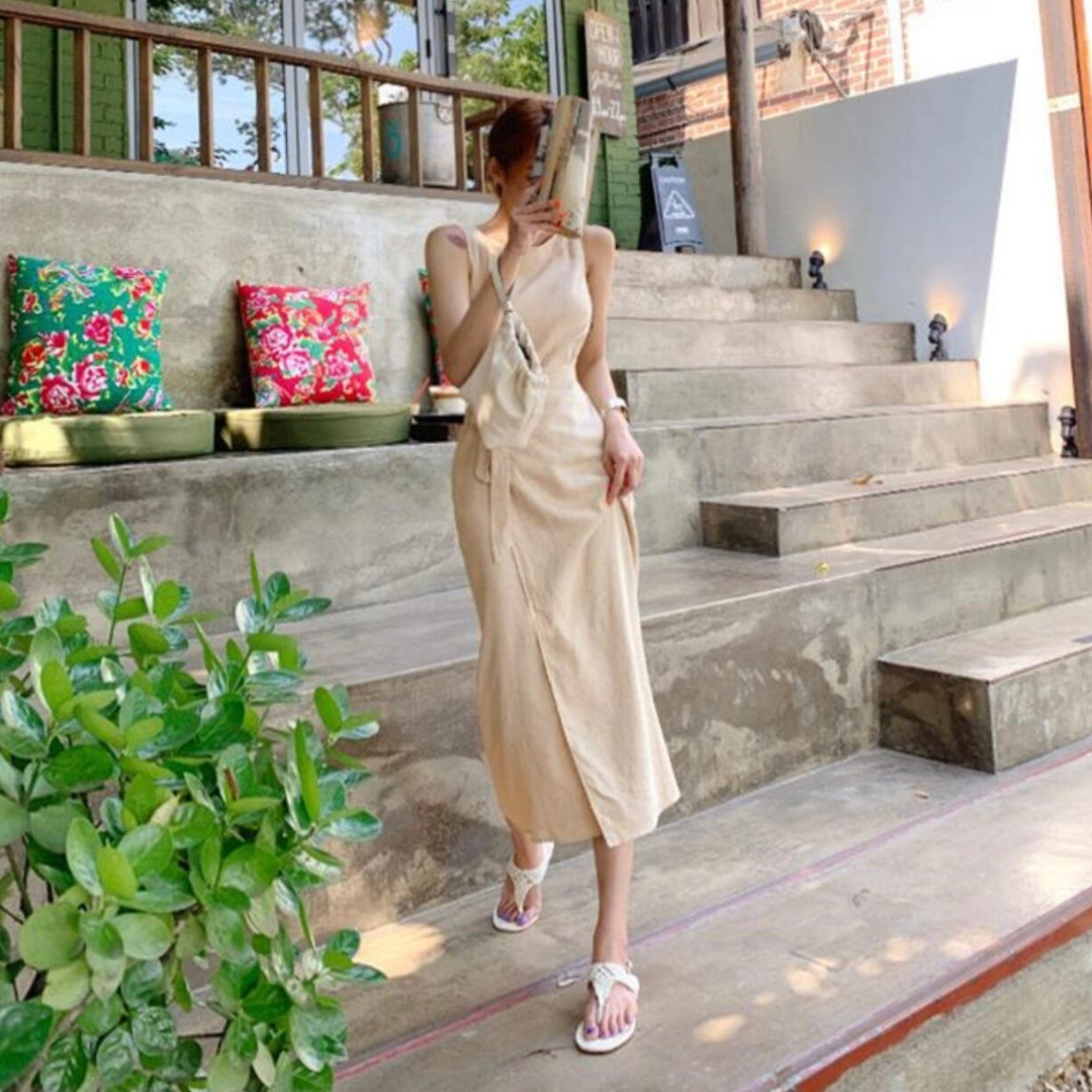 Frau in beigem Sommerkleid auf Treppe, umgeben von Pflanzen und bunten Kissen. Outdoor-Mode, lässiger Stil, Sommeroutfit.
