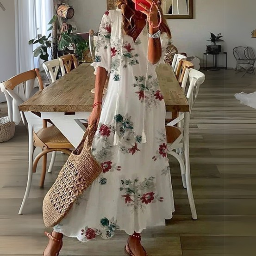 Frau in langem, geblümtem Sommerkleid mit Strohtasche steht in modernem Esszimmer. Holztisch, Stühle, Pflanzen im Hintergrund.