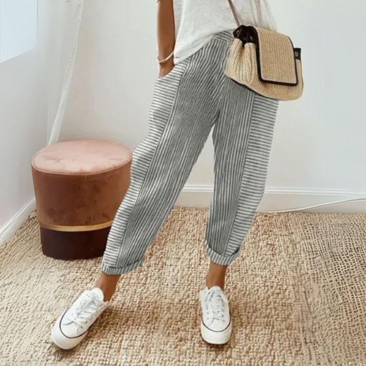 Frau in gestreifter Hose und weißen Sneakers, hält eine Strohtasche. Moderner, lässiger Look, ideal für Sommermode und Freizeitkleidung.