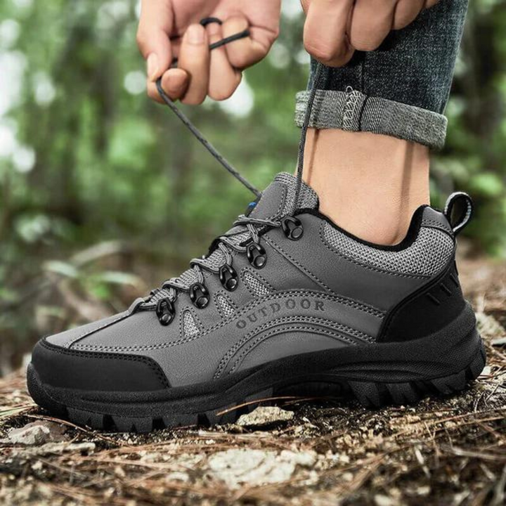 Grauer Outdoor-Wanderschuh mit robustem Profil, geschnürt auf Waldboden. Ideal für Trekking und Naturabenteuer. Hochwertige Materialien.