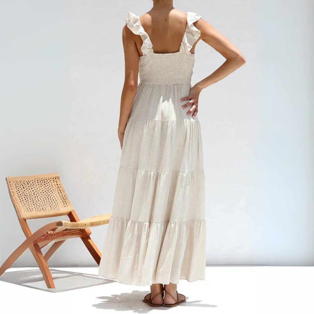 Rückenansicht einer Frau in einem langen, weißen Sommerkleid mit Rüschen, neben einem modernen Holzstuhl. Perfekt für Sommermode und Strandkleidung.