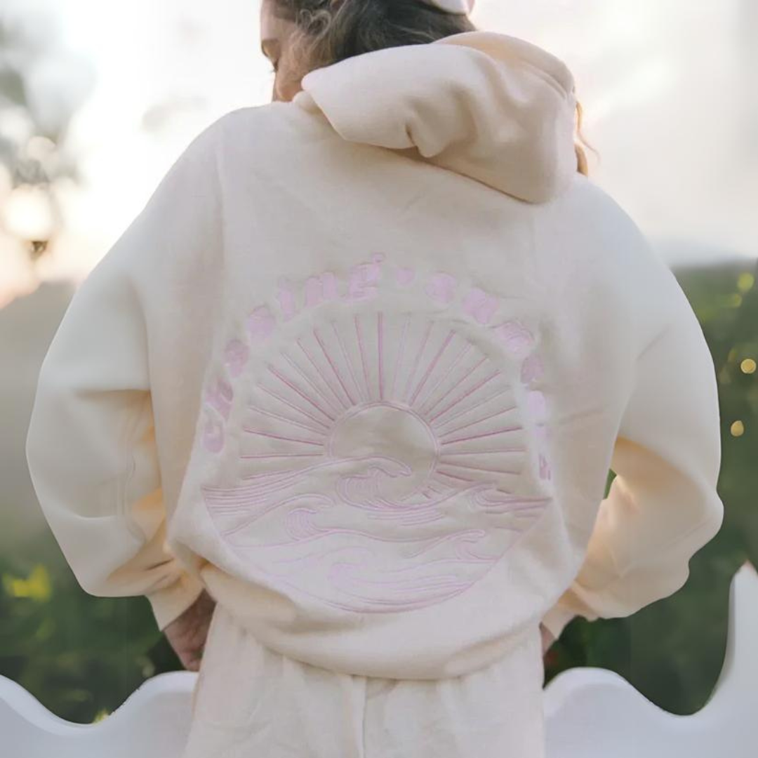 Person in cremefarbenem Hoodie mit Sonnenaufgangs- und Wellenmotiv auf dem Rücken, im Freien bei Sonnenuntergang. Bequeme Freizeitmode, Natur.