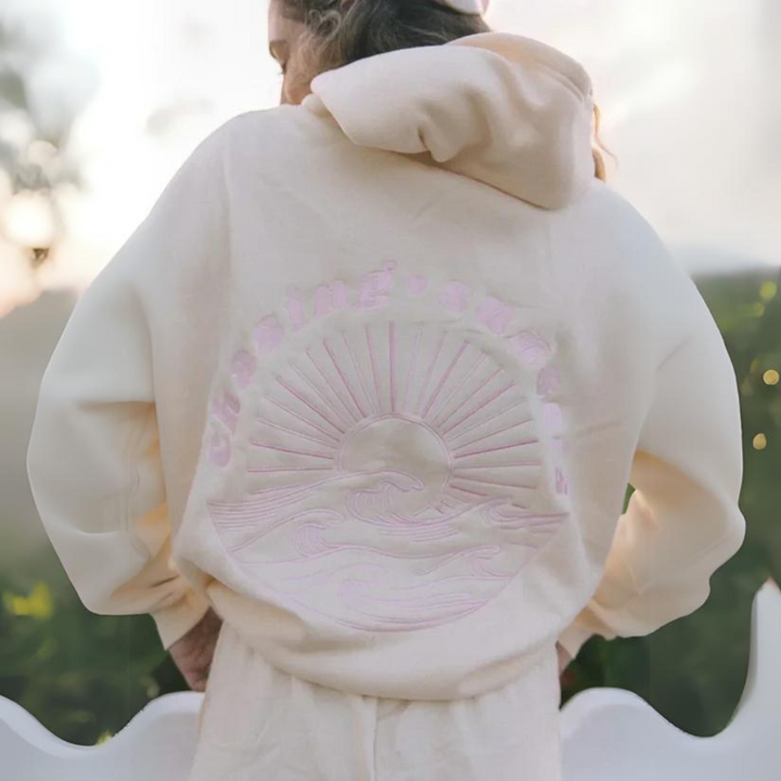 Person in cremefarbenem Hoodie mit Sonnenaufgangs- und Wellenmotiv auf dem Rücken, im Freien bei Sonnenuntergang. Bequeme Freizeitmode, Natur.
