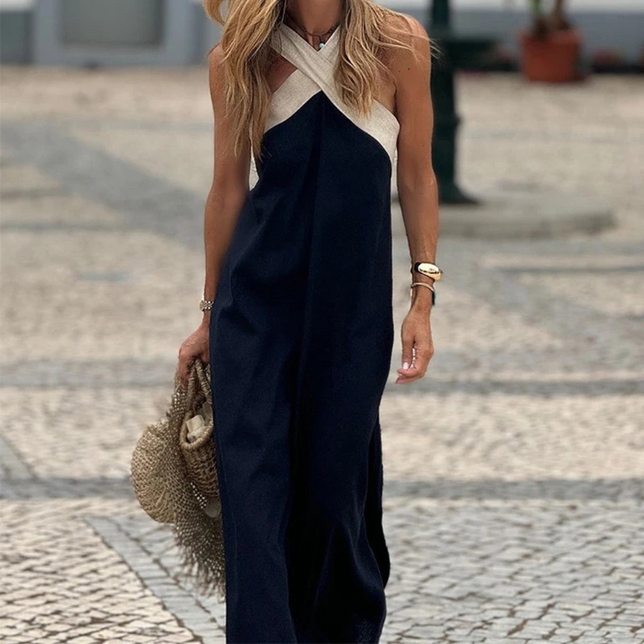 Frau in elegantem, ärmellosem Maxikleid mit überkreuztem Ausschnitt, hält Strohtasche. Sommermode, stilvoll, urbaner Hintergrund.