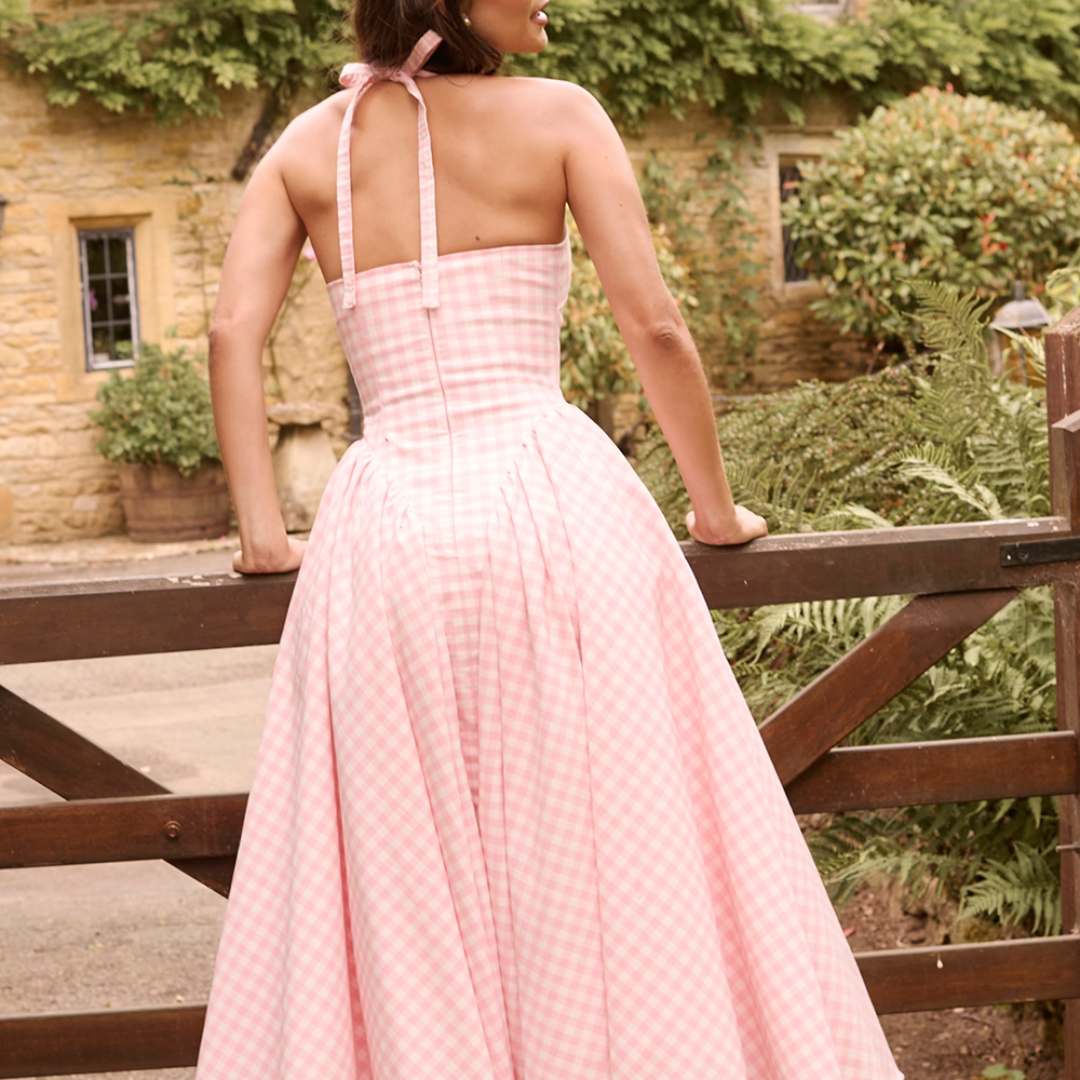 Rückansicht einer Frau in einem rosa karierten Sommerkleid vor einem rustikalen Holztor, umgeben von grüner Vegetation und einem Steinhaus.