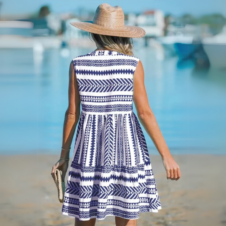 Frau in blau-weiß gemustertem Sommerkleid und Strohhut am Strand, hält eine Clutch. Perfektes Outfit für Strandurlaub und Sommermode.