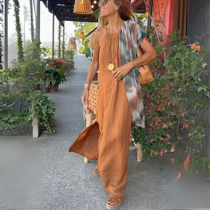 Frau in langem, orangefarbenem Kleid und Sandalen spaziert auf Gehweg, trägt goldene Halskette und braune Tasche, umgeben von grünen Pflanzen.