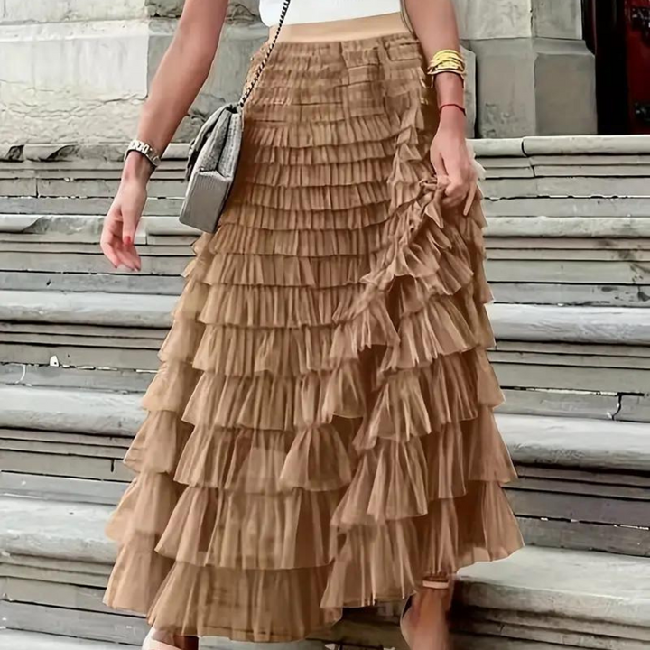 Frau mit braunem, plissiertem Rüschenrock auf Treppe, trägt weiße Bluse, graue Handtasche und goldene Armbänder. Modetrend, eleganter Look.