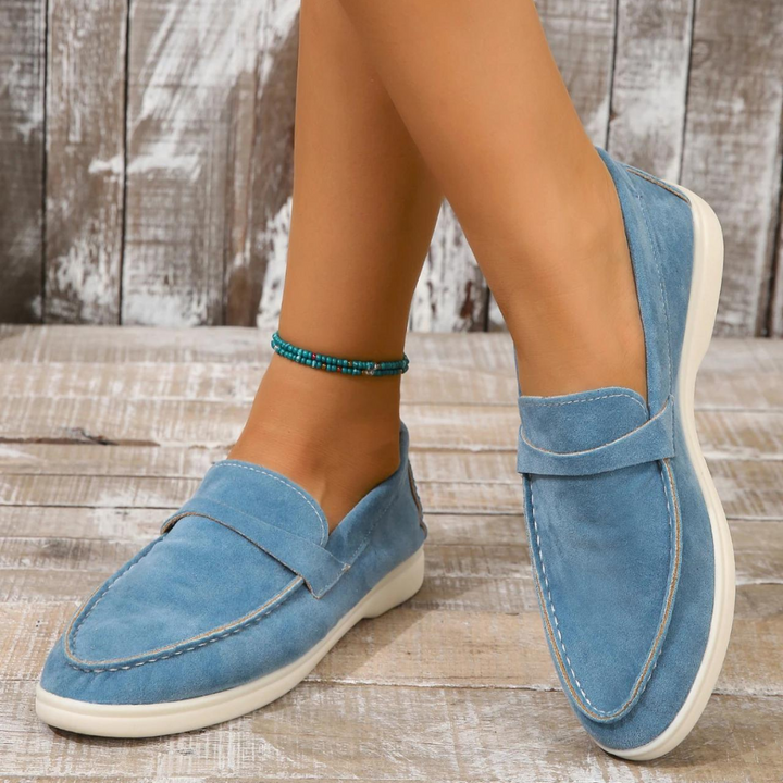 Blaue Wildleder-Loafer für Damen auf Holzboden, getragen mit türkisfarbenem Fußkettchen. Bequeme, stilvolle Schuhe, ideal für Freizeitmode.