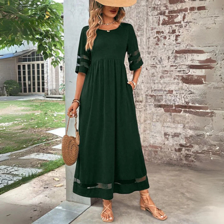 Frau in elegantem, langem, grünem Kleid mit Strohhut und runder Strohtasche, steht vor einer Backsteinmauer. Sommermode, Damenkleid, Outdoor.