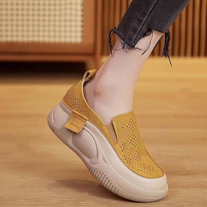 Gelber Damen-Sneaker mit dicker Sohle, perforiertem Design und elastischem Einsatz, getragen auf Holzfußboden. Modischer Freizeitschuh für Damen.