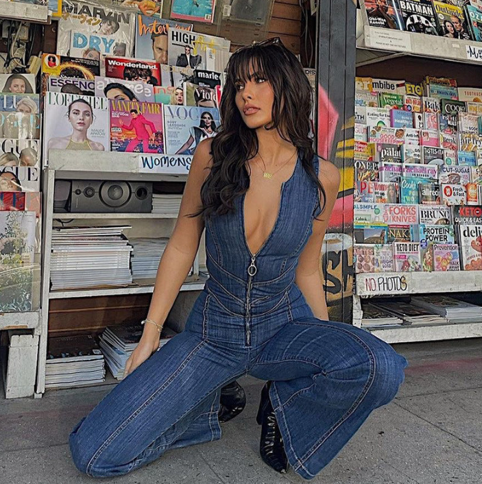 Frau in blauem Jeans-Overall posiert vor einem Zeitschriftenstand. Mode, Streetstyle, trendige Kleidung, Zeitschriften im Hintergrund.