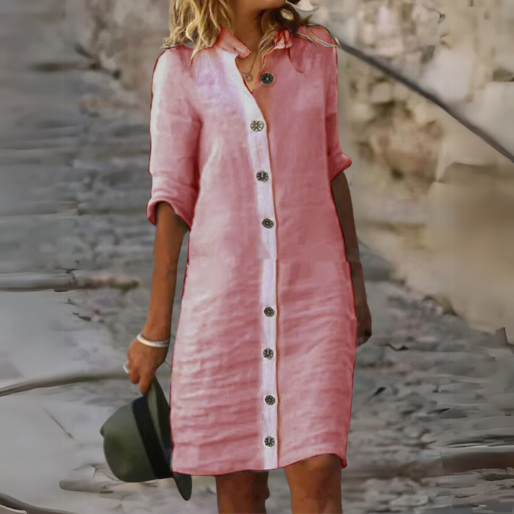 Frau in rosa Leinenkleid mit Knopfleiste, lässig auf Kopfsteinpflaster gehend, hält Hut in der Hand. Sommermode, Freizeitkleidung, Damenkleid.