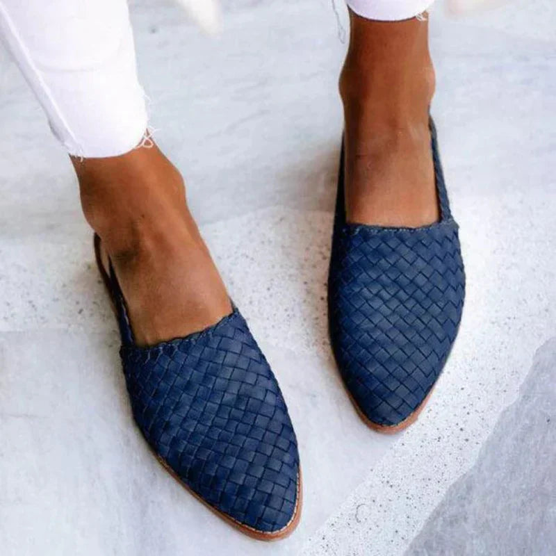 Damenfüße in blauen, geflochtenen Slippern auf grauem Boden, weiße Jeans. Modische Sommerschuhe, bequeme Damen-Slipper, trendige Fußbekleidung.
