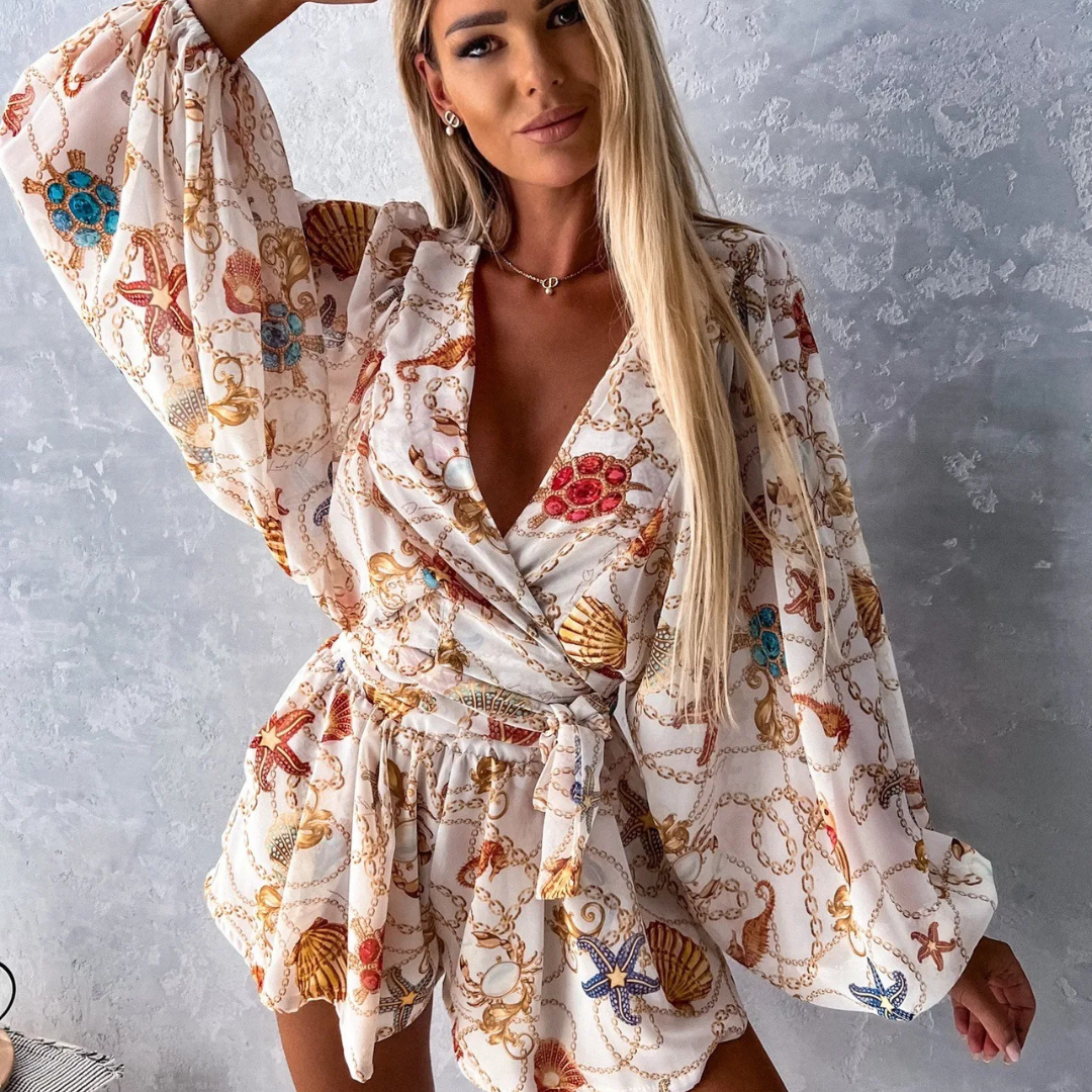 Frau in elegantem, maritimem Jumpsuit mit Muschel- und Kettenmuster vor grauer Wand. Modetrend, Sommeroutfit, Damenmode.