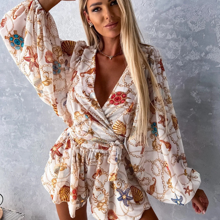 Frau in elegantem, maritimem Jumpsuit mit Muschel- und Kettenmuster vor grauer Wand. Modetrend, Sommeroutfit, Damenmode.