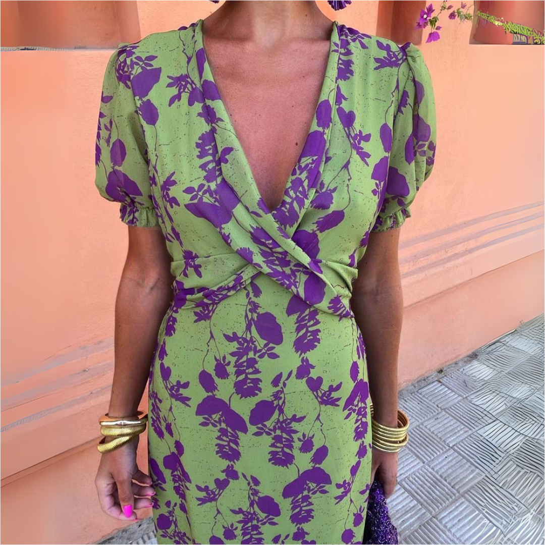 Frau in grünem Kleid mit lila Blumenmuster, tiefem V-Ausschnitt und Puffärmeln. Goldene Armreifen, sommerlicher Look, modisch, elegant.