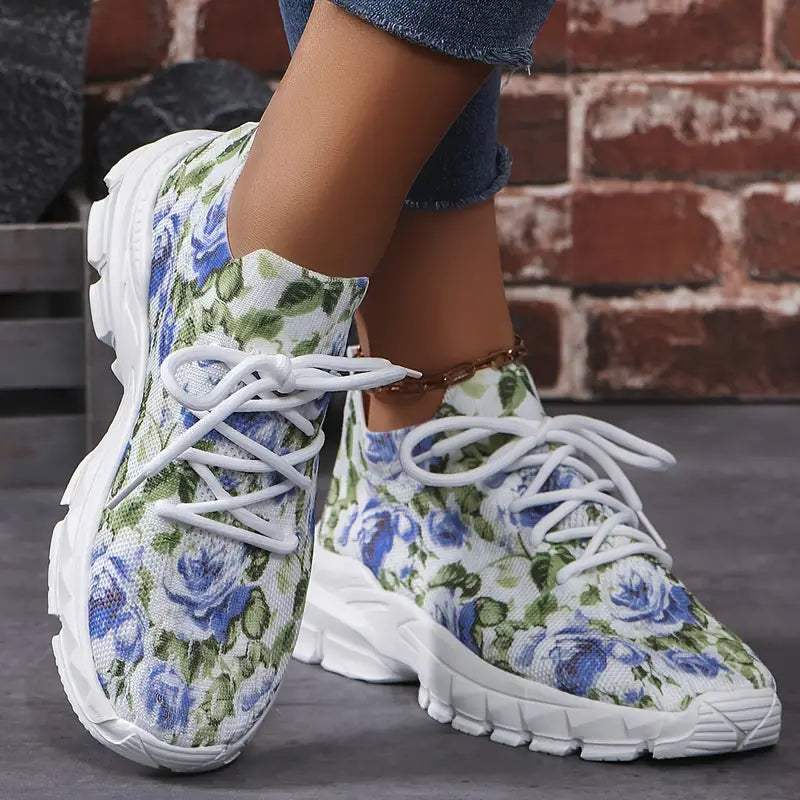 Damen-Sneaker mit blau-grünem Blumenmuster, weiße Schnürsenkel, bequeme Sohle, ideal für Freizeitmode und Frühlingsoutfits vor Ziegelwand.