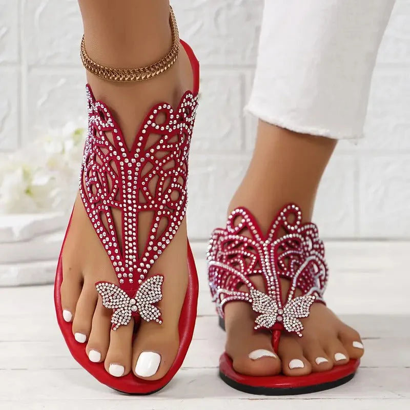 Rote Sandalen mit Strassverzierung und Schmetterlingsdesign, getragen von einer Person mit weiß lackierten Zehennägeln auf hellem Hintergrund.