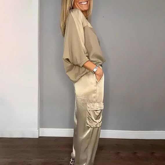 Frau in beiger Bluse und glänzender Satin-Cargohose, lächelnd, seitlich stehend vor grauer Wand. Modetrend, lässiger Stil, elegante Damenmode.
