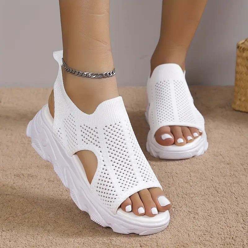 Weiße Damen-Sneaker-Sandalen mit Strickmuster und dicker Sohle, getragen an Füßen mit weiß lackierten Nägeln. Modische Sommerschuhe für Frauen.