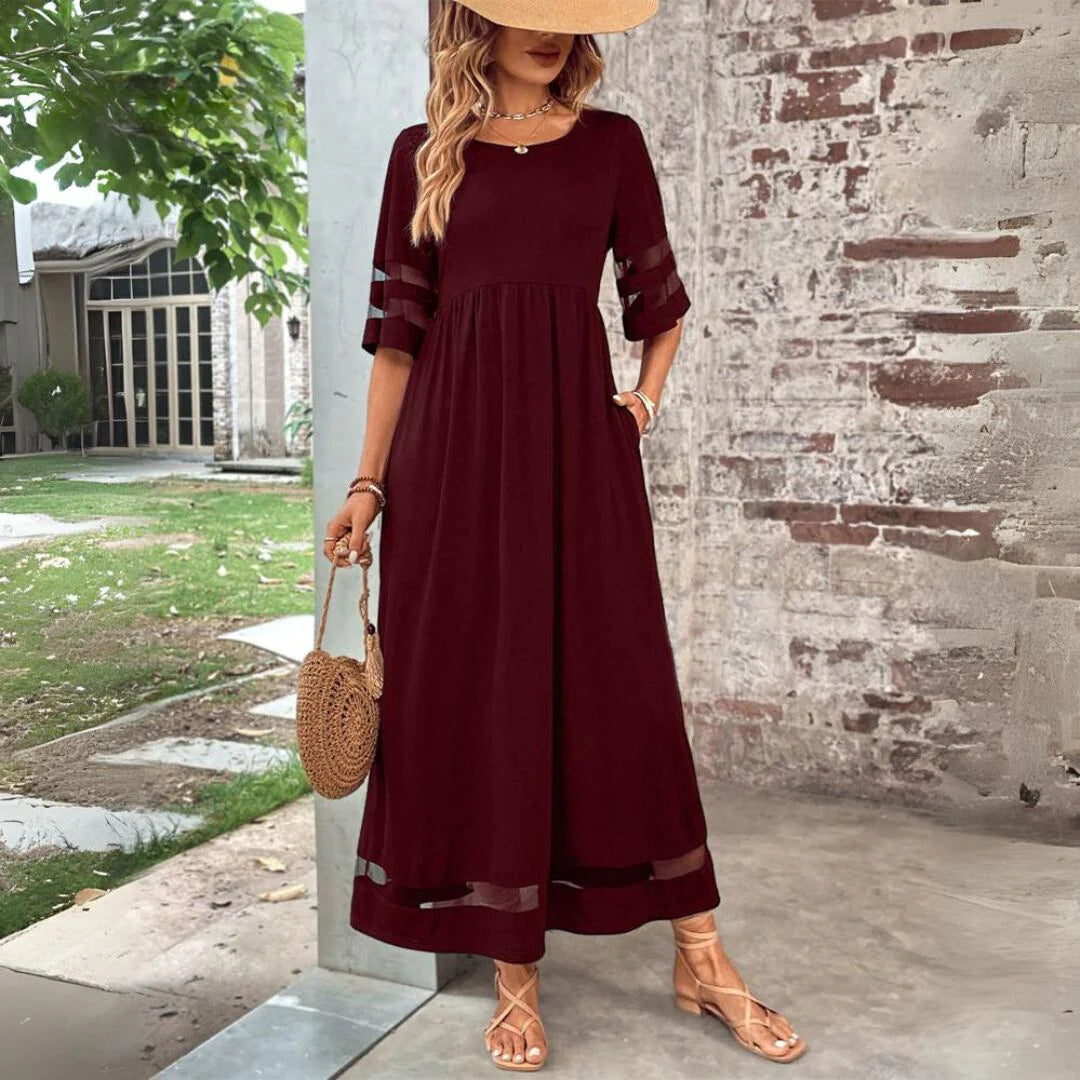 Frau in elegantem, bordeauxrotem Maxikleid mit transparenten Ärmeln, Strohhut und Sandalen, steht vor einer Backsteinmauer. Sommermode, Boho-Stil.