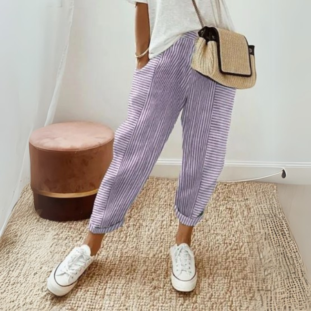 Frau in gestreifter Hose und weißem T-Shirt, trägt eine Strohtasche und weiße Sneaker, steht auf einem Teppich neben einem rosa Hocker.