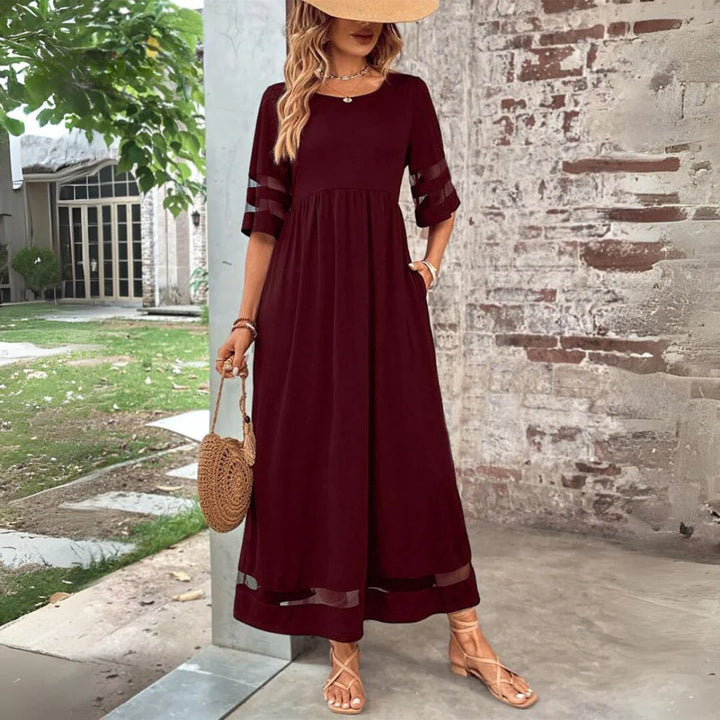 Frau in elegantem, bordeauxrotem Maxikleid mit transparenten Ärmeln, Strohhut und runder Strohtasche, steht vor einer Backsteinmauer im Freien.