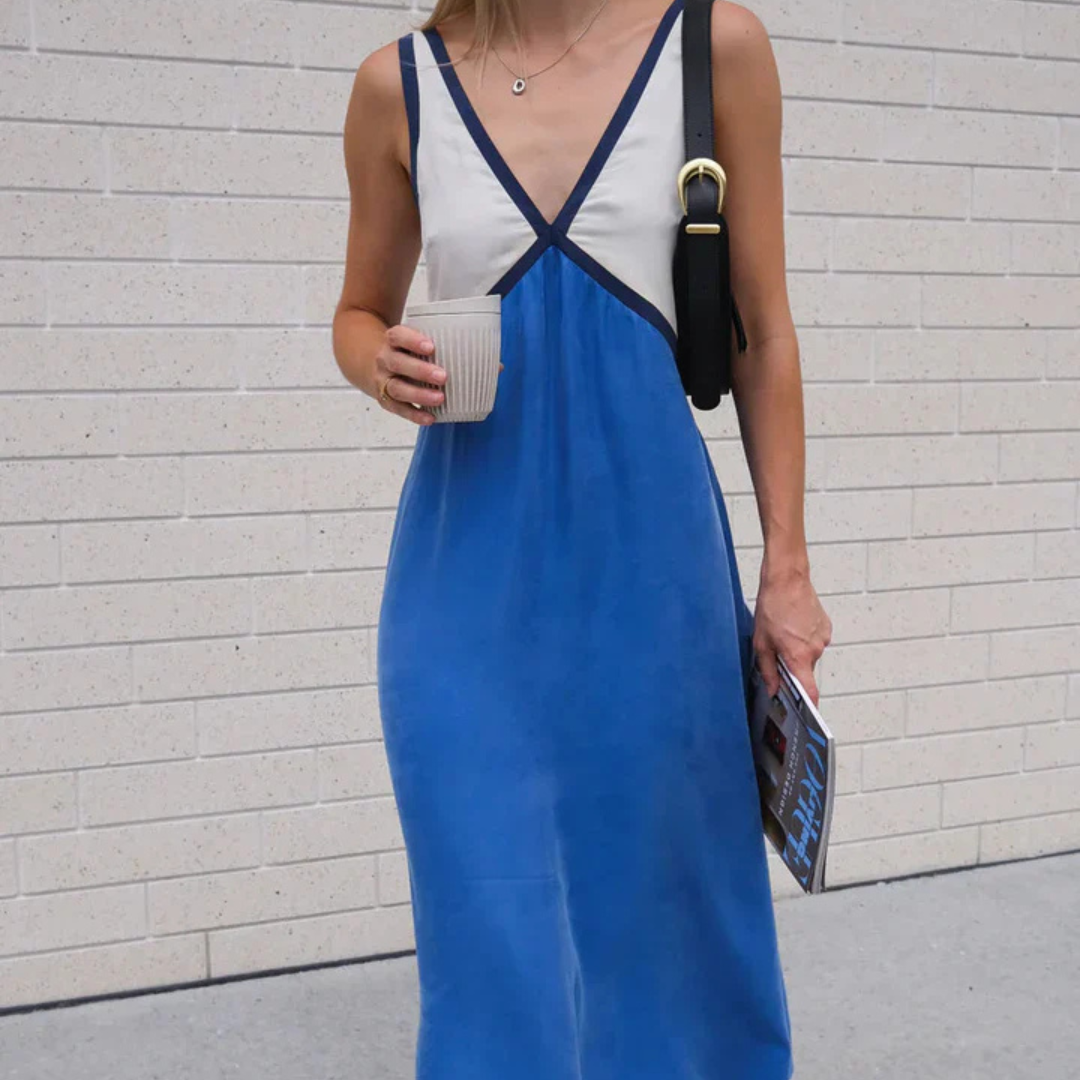 Frau in blau-weißem Sommerkleid mit Kaffeebecher und Zeitschrift vor heller Ziegelwand. Modetrend, Sommeroutfit, Freizeitkleidung.
