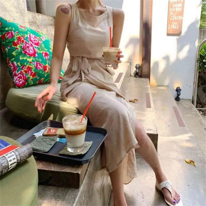 Eine Frau in einem beigen Kleid sitzt auf einer Terrasse und hält ein Getränk. Auf dem Tisch stehen Eiskaffee und Zeitschriften. Entspannte Sommeratmosphäre.