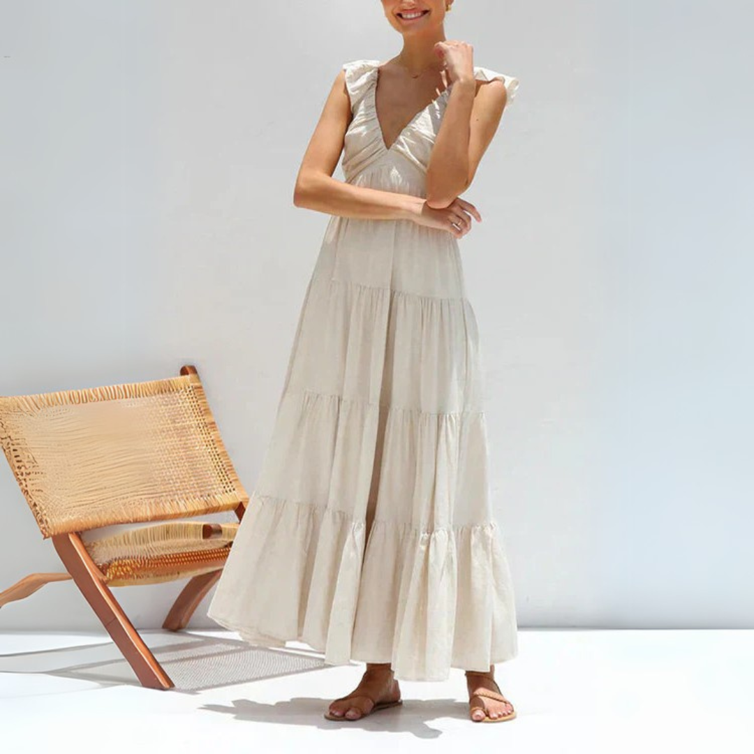 Frau in weißem Sommerkleid mit Rüschenärmeln, steht neben einem modernen Holzstuhl. Elegantes, luftiges Maxikleid für warme Tage.