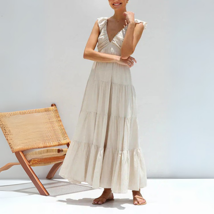 Frau in weißem Sommerkleid mit Rüschenärmeln, steht neben einem modernen Holzstuhl. Elegantes, luftiges Maxikleid für warme Tage.