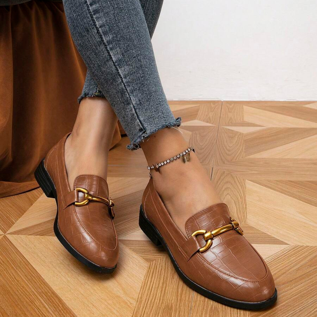 Braune Damen-Loafer mit goldener Schnalle, getragen mit grauen Jeans auf Parkettboden. Modische Schuhe, elegantes Design, ideal für Herbstmode.
