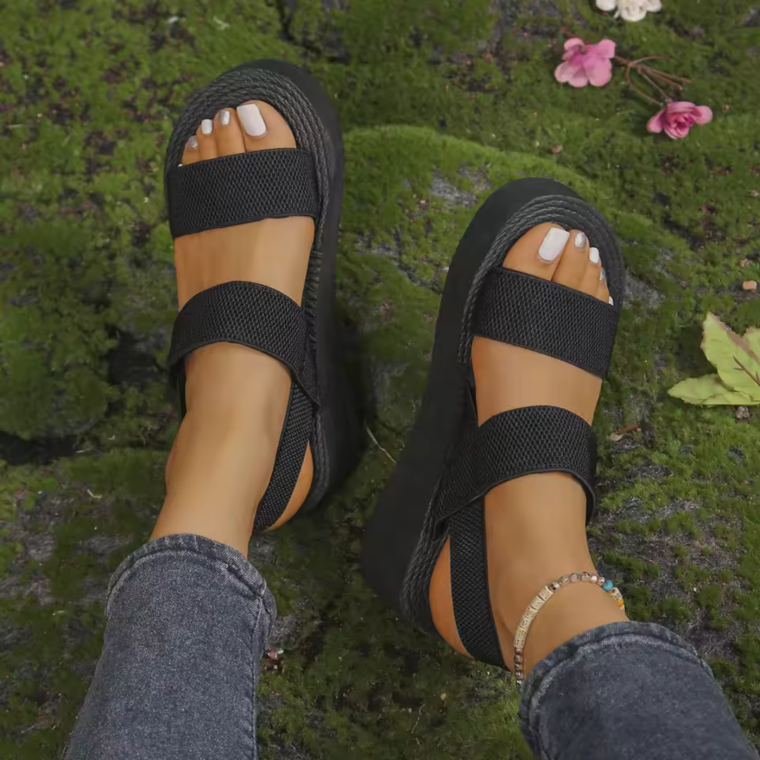 Schwarze Plateau-Sandalen mit Klettverschluss an Frauenfüßen, getragen auf moosbedecktem Boden. Modische Sommerschuhe, bequem und stilvoll.