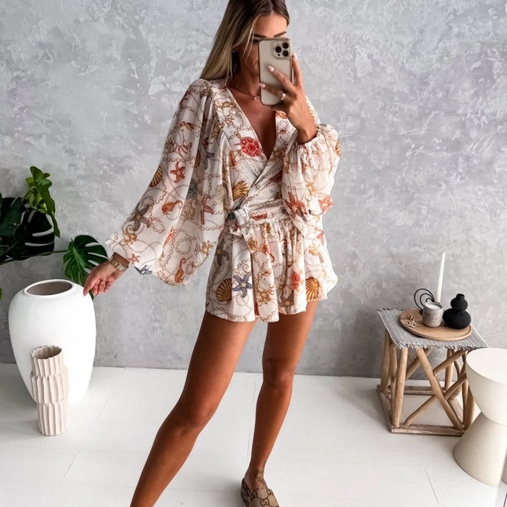 Frau in geblümtem Sommerkleid posiert vor grauer Wand, hält Smartphone. Moderne Inneneinrichtung mit Pflanzen und Dekoration im Hintergrund.