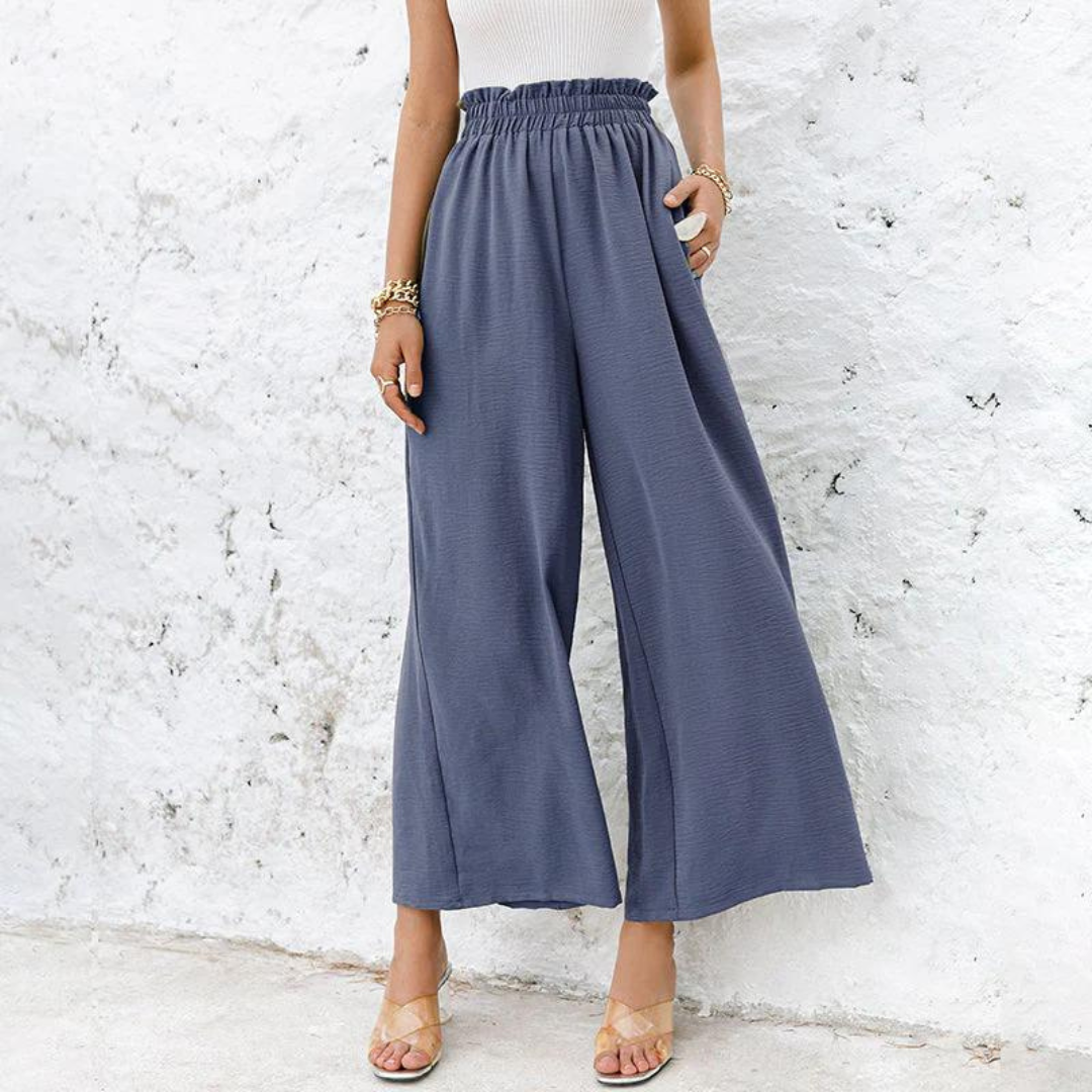 Frau trägt weite, blaue Palazzo-Hose mit hoher Taille, kombiniert mit Sandalen und Armbändern, vor weißer Wand. Modetrend, Damenbekleidung.