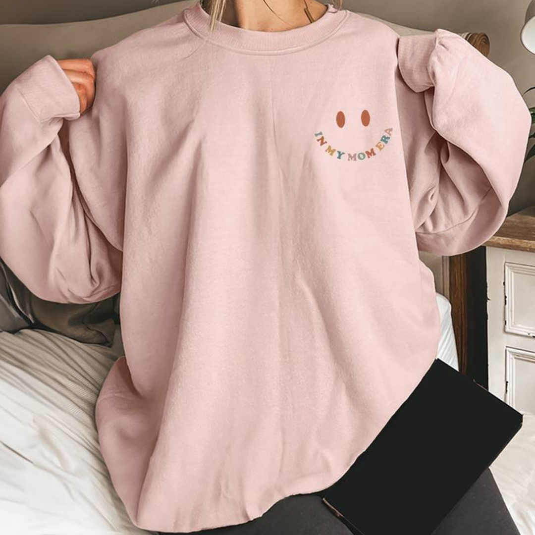 Frau trägt oversized rosa Sweatshirt mit Smiley-Design und "Not My Momma" Aufschrift, sitzt auf Bett. Bequemes, modisches Freizeitoutfit.