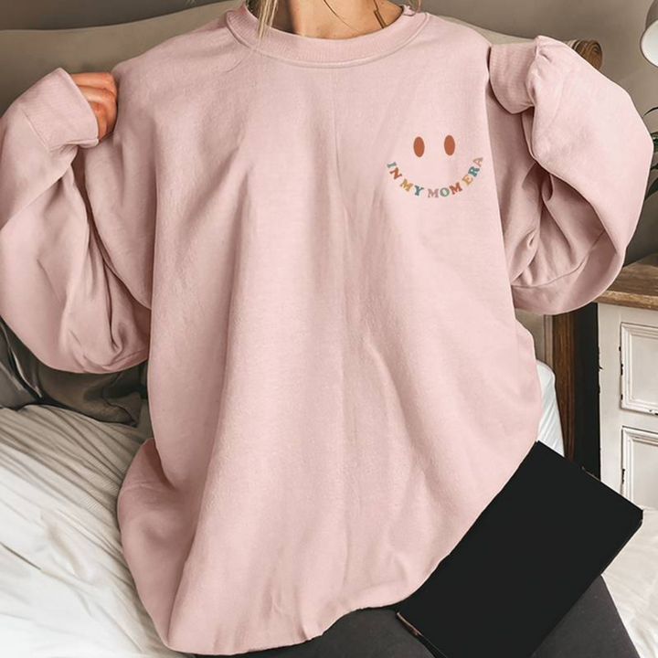 Frau trägt oversized rosa Sweatshirt mit Smiley-Design und "Not My Momma" Aufschrift, sitzt auf Bett. Bequemes, modisches Freizeitoutfit.